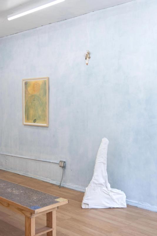 a view of a well lit gallery space with a cloudy light blue wall and vinyl wood flooring. in the foreground is a large bench. in the center is a white sculpture on the floor covered in white fabric, and loosely in the shape of a very skinny tall mountain. on the wall is a tellow framed artwork and a small sculpture thats not visible in detail.