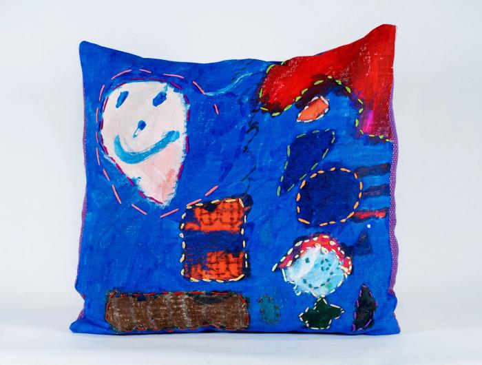 a blue rectangular pillow. on the top left is a white face in a balloonish shape with blue smile. bottom right is another figure and they are surrounded by other shapes, a small orange square and a long brown rectrangle.