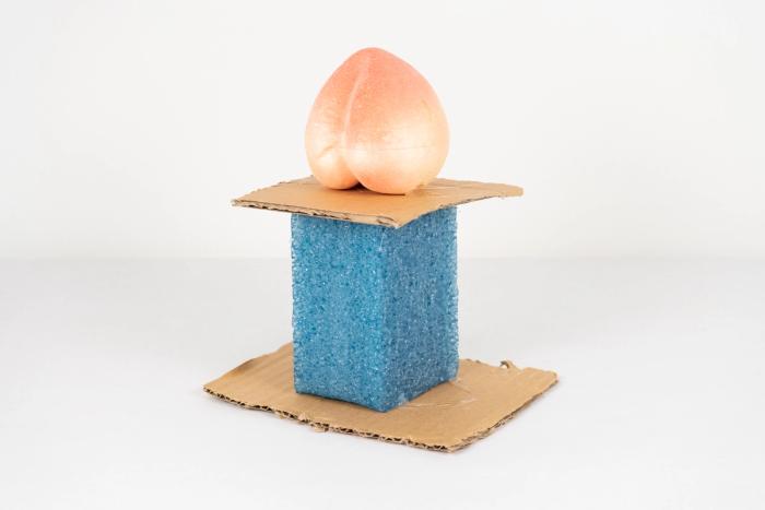A cardboard and styrofoam sculpture of a pedestal with a large peach shaped object on top.