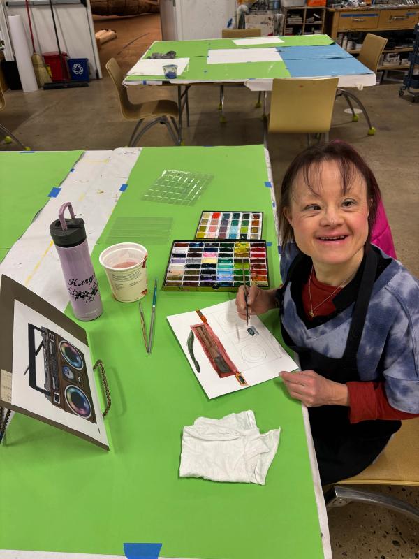 Portrait of the artist working on a painting of a boombox at a bright green studio table, surrounded by brushes and other art supplies.