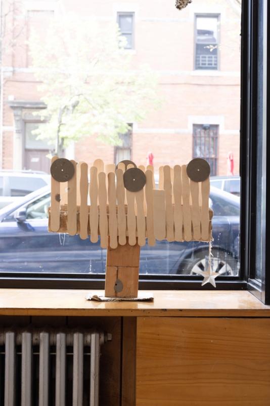 a sculpture standing on a built in window bench in front of a window looking out on a street on a bright day. the sculpure stands about a foot tall and two wide and has a wooden base, clean popsicle sticks upright in a loose fence-like formation and three evenly spaced circular forms on the top. hanging off the right side is a silver star. 