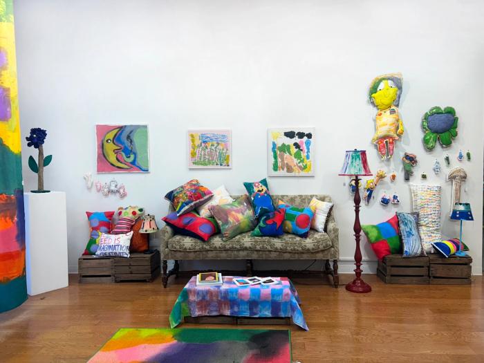 a brightly lit gallery space with vividly colored objects. on the wall are three paintings, one discernible as a moon smoking a pipe. there is a small coffee table with a checkered table cloth and books, a couch with bright cushions and a lamp. at left is a small sculpture of a purple flower on a pedestal.