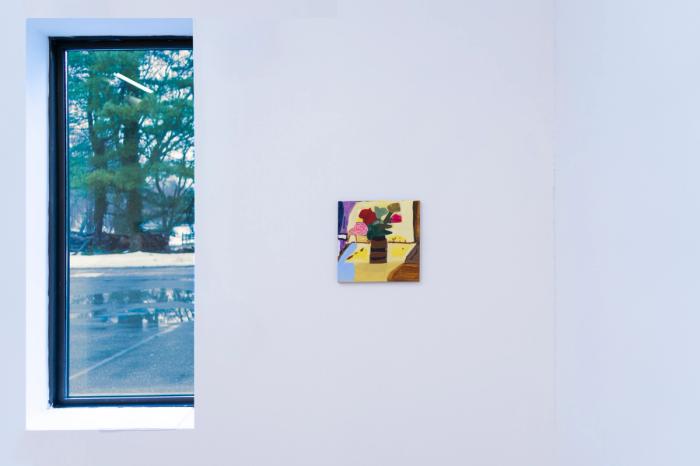 a small square shaped painting on a white gallery wall. to its left is a window on a parking lot with a tree. the painting is mostly yellow and is of a vase with flowers in it. 