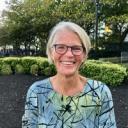 a woman with pale skin and a silver bob wears glasses and smiles towards the camera. she is standing outside in front of some landscaped bushes with trees in the background