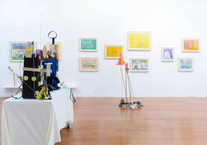 a white walled gallery space with wooden floor. there are lots of artworks in bright colors in light wooden frames on the wall. in the foreground are two sculptures, one is a very intricate box shape with lots of things hanging off, the other is a standing structure with a pink shape in the top.