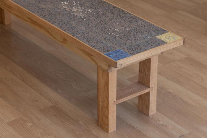 a wooden framed bench with a very intricate pebbly cement-like top section thats inset into the wooden frame. most of the top is dark gray brown with each corner containing a small square of a different color, red, blue/green, yellow, dark blue. 