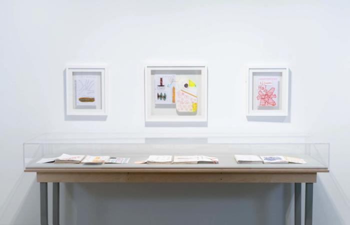 a view of a gallery wall with three framed artworks on the wall. they are multicolored and the one in the middle is an organic shape. there is a vitrine table in the foreground with books laid flat inside, pages open.