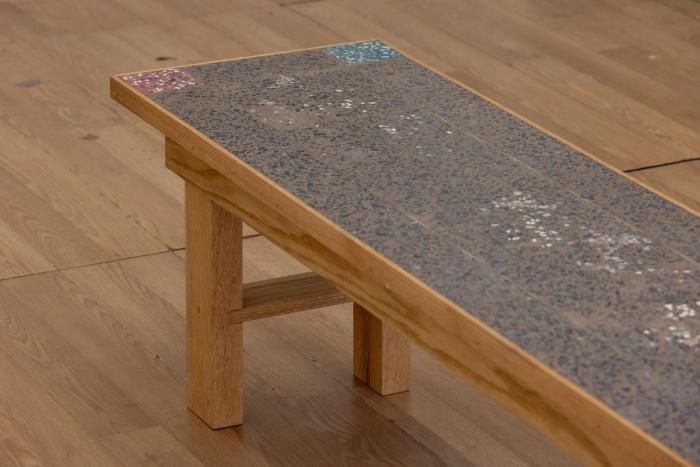 a wooden framed bench with a very intricate pebbly cement-like top section thats inset into the wooden frame. most of the top is dark gray brown with each corner containing a small square of a different color, red, blue/green, yellow, dark blue. 