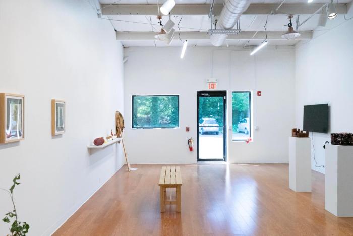 a view of an art gallery with white walls, sculpures on white plinths, and several images in paintings on the wall. theres a monitor in the back left and a shelf with sculptures