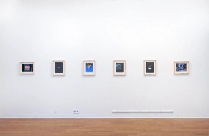 a white walled gallery with wooden floor and artworks in pale wooden frames on the wall in a single line. they are multicolored.