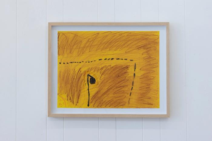a framed artwork on a white bead board wall. the drawing is on yellow paper and focuses on a dark brown/black form that appears like a streetlight. going from left and turning down to the bottom middle is a dotted line that appears like a road or path.