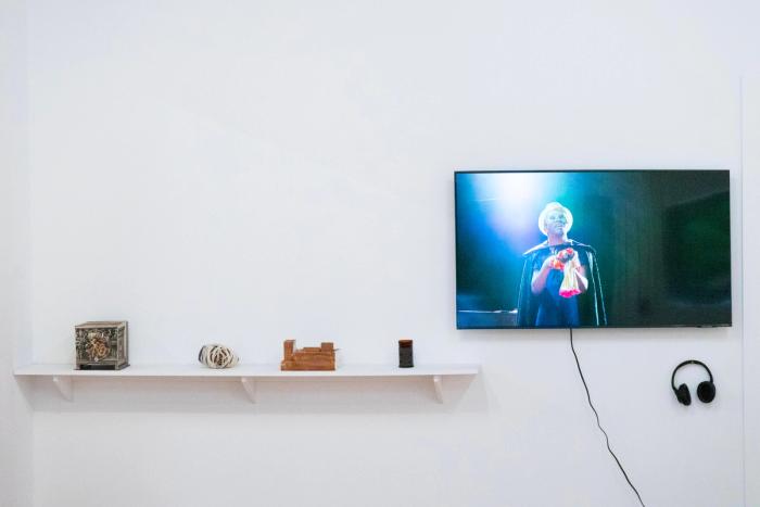 several small scultpures are installed on a white shelf, above them and to the right is a monitor displaying an image of a man holding a puppet. 