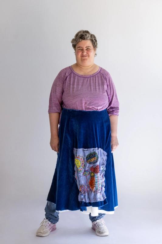 The artist standing against a white wall modeling her blue apron with custom embroidery, a lavender top, and blue jeans.