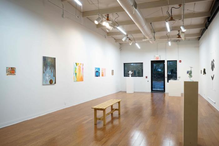 a view of a gallery. there are two dimensional artworks on the walls and a pedestal with a sculpture in the foreground. the viewpoint is towards the front door and windows. the room is white and brightly lit.