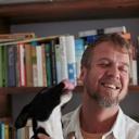 a white man with greying blond facial hair smiles away from the licking face of a small black and white dog. there are lots of books on shelves in the background