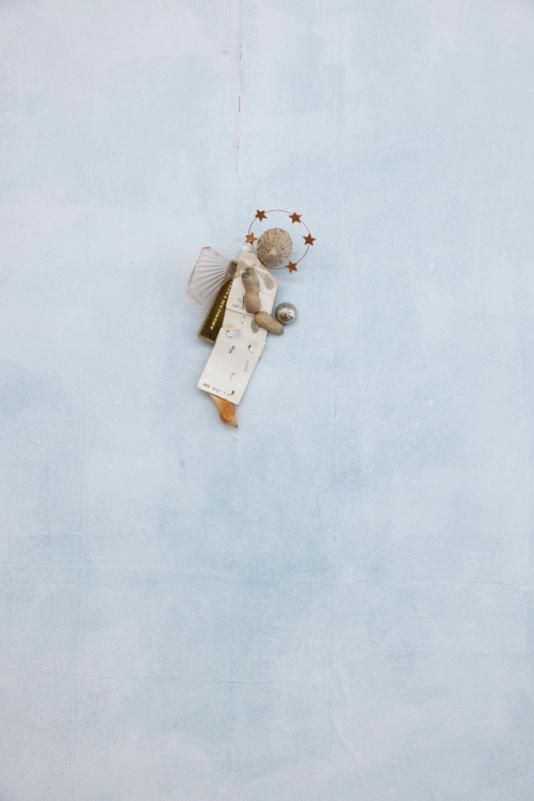 a sculpture in the shape of a figure against a cloudy blue/gray wall. The figures body is made of a piece of cardboard from some priduct, a shell as a head, arms made of peanuts, a foot made of a small shell, starry halo, and holding a chrome marble.