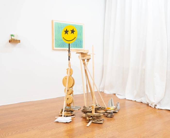 a corner of a white walled gallery with a sculpture int he center of the frame. its made of wood scraps and has a big yellow smiley face with stars for eyes on the top of a spire