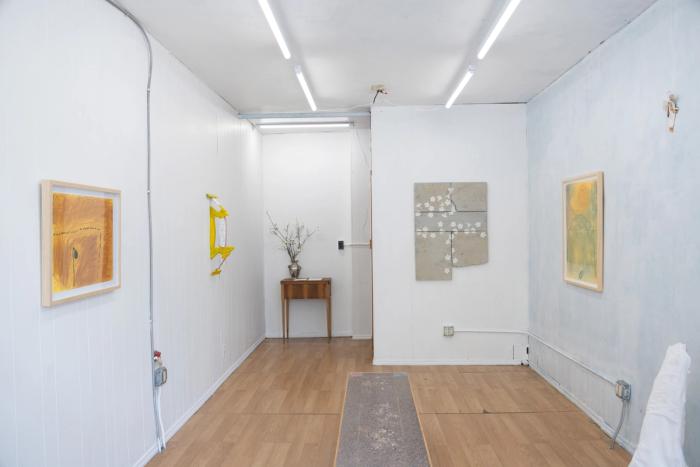 a view of a small well lit gallery space with vinyl wooden floors and white walls, lit by tube lights. there are five artworks on the walls, two are framed drawings, and one is made of several distinct cast conrete pieces that meet and make a roughly rectangluar shape