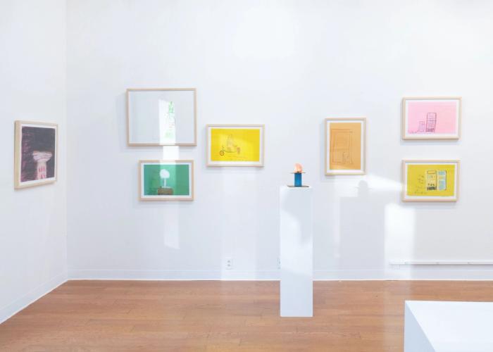 a white gallery wall with cool light coming in from windows outside the frame making bright spots on the wall. there are 8 artworks in frames hanging, several on brightly colored paper. in the foreground is a pedestal with a small sculpure of a peach on a tiny blue pedestal 