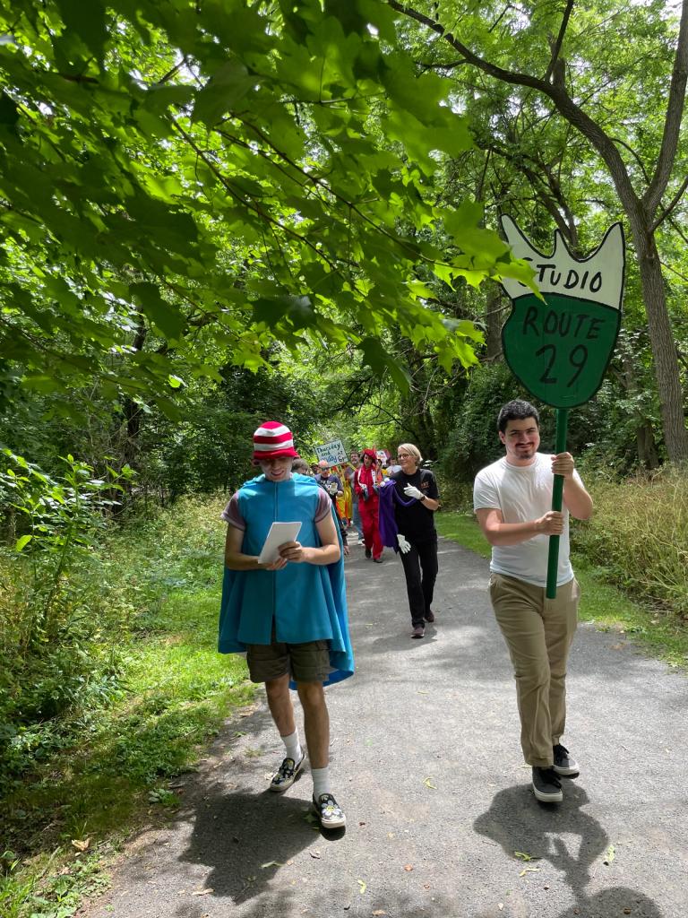 people walking on a forested path wearing costumes. One holds a sign in the shape of a a badge with writing that says 'studio route 29'