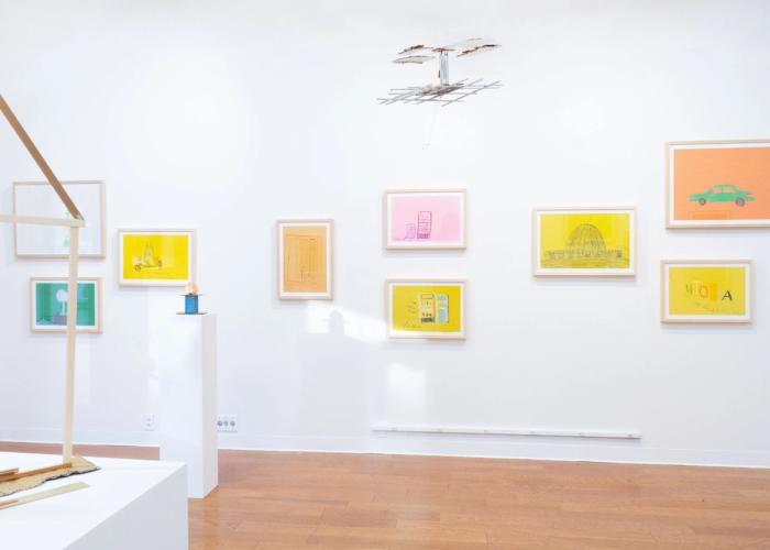 a white gallery wall with a wooden floor. there are nine artworks in frames hanging on the wall in various bright colors, mostly warm ones. hanging above from the ceiling is a silver sculpture. against the wall is a pedestal with a small sculpture with a blue form