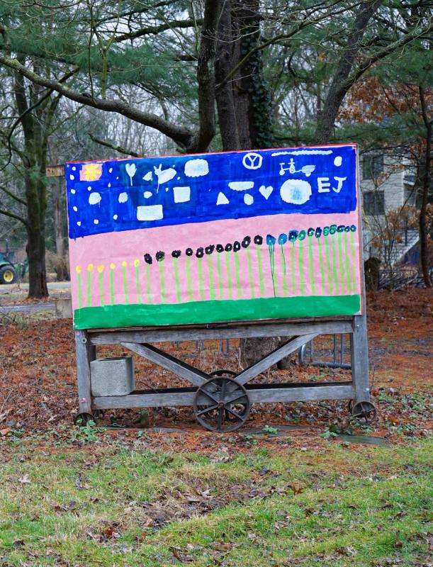 a sign is painted on a wheeled wooden cart thats set outside with trees and grass. the painting is of a row of tall straight flowers and a blue sky with clouds in all kinds of shapes even a heart and a firetruck