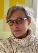 a white woman with blond hair pinned back smiles with lips together into the camera. shes wearing red and brown glasses and a heavy wool sweater.