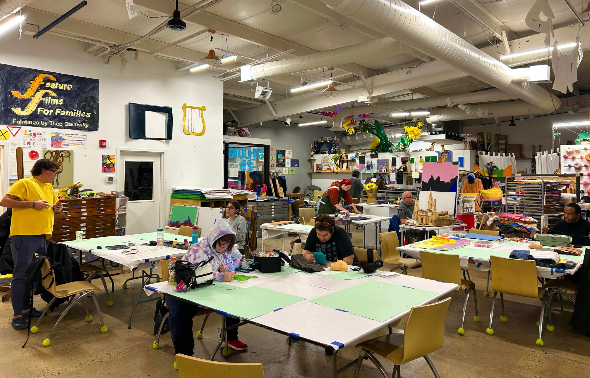 a bright and colorful room with artists working and some talking. tables are spread out through the space which has a tan concrete floor and white walls with artworks hanging.