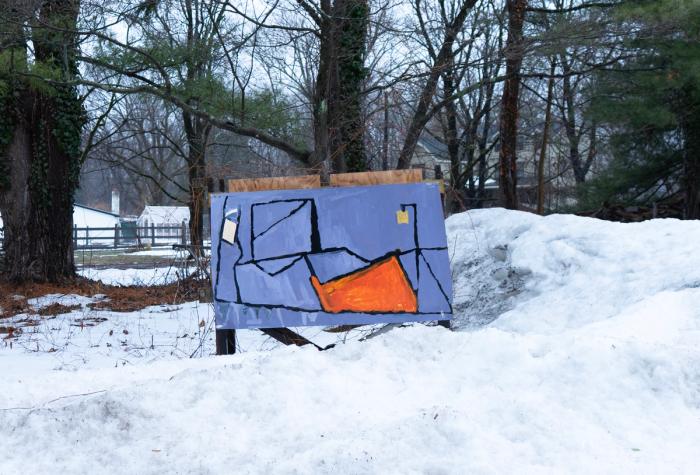 a painting installed outside on a cart. Theres snow in the fore and middle ground and trees in the background. a few houses are visible in the background as well. The painting is mostly gray and has dark black lines running in loose rectangles with one big orange section and a small white and small yellow section