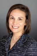 A white woman with a straight brown bob smiles at the camera. she wears a string of pearls and a rough woven sport coat.