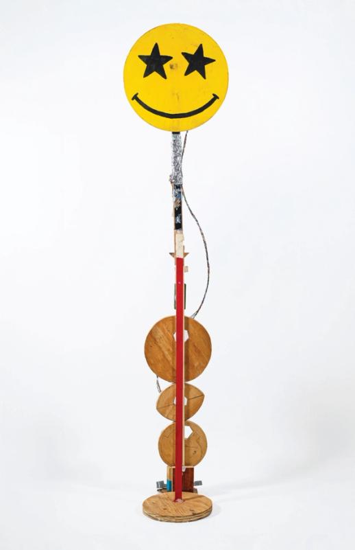 a sculpture in a white space with a slight shadow around the base. its mostly made of wood and is thin and tall. at the bottom are four loosely round pieces of wood in different shapes. the central form is a thin piece of wood partially painted red. at the top is a big yellow smiley face with star eyes. there is reflective foil beneath and a wire going down to the base.