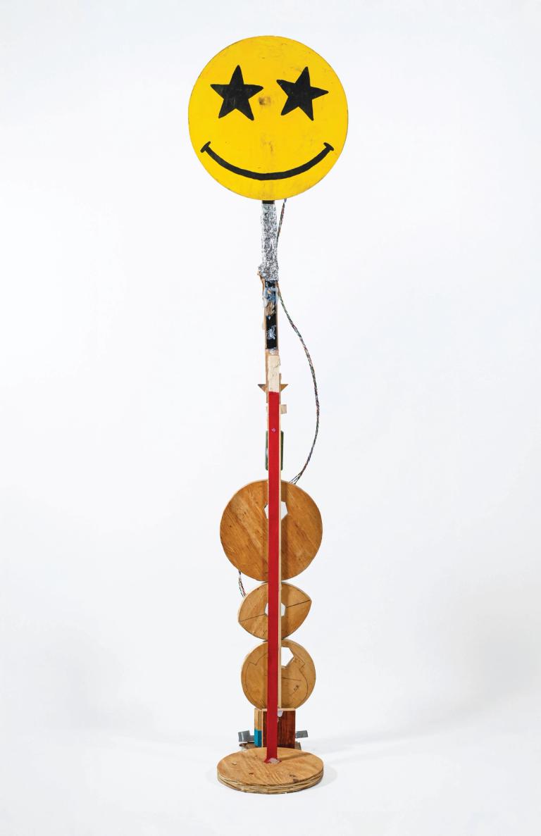 a sculpture in a white space with a slight shadow around the base. its mostly made of wood and is thin and tall. at the bottom are four loosely round pieces of wood in different shapes. the central form is a thin piece of wood partially painted red. at the top is a big yellow smiley face with star eyes. there is reflective foil beneath and a wire going down to the base.
