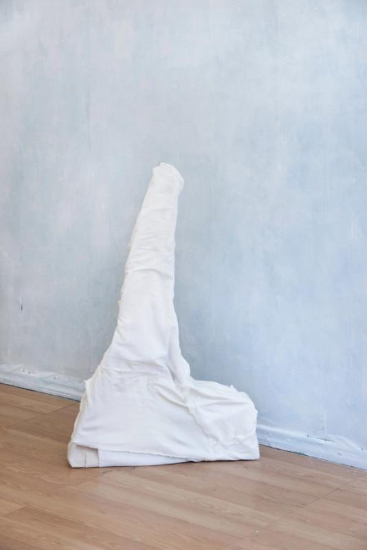 a white sculpture on a vinyl wooden floor against a foggy blue/gray wall. the sculpture is covered in pieces of white textile and has a broad solid shaped base section and a thinner, tall upright section.