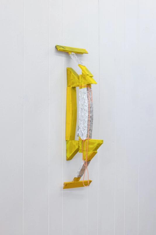 a yellow sculpture installed on a white bead board wall. its mostly yellow and roughly symmetrical. the center section is wrapped in aluminum foil and it loosely takes the shape of a bridge. it has an orange string attached to it and is intalled with the bottom of the bridge against the wall