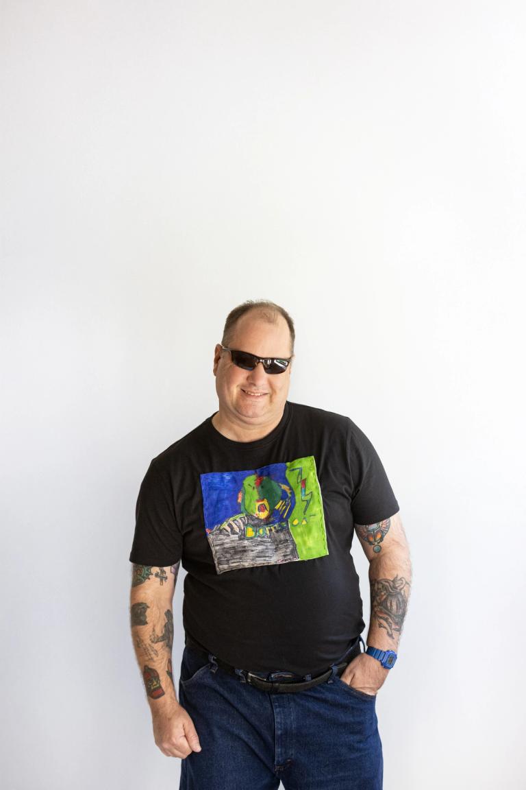 a light skinned man with dark sun glasses and many arm tattoos smiles at the camera. Hes wearing a black shirt with a colorful patch attached to the front. He looks happy and casual with one hand in his pocket