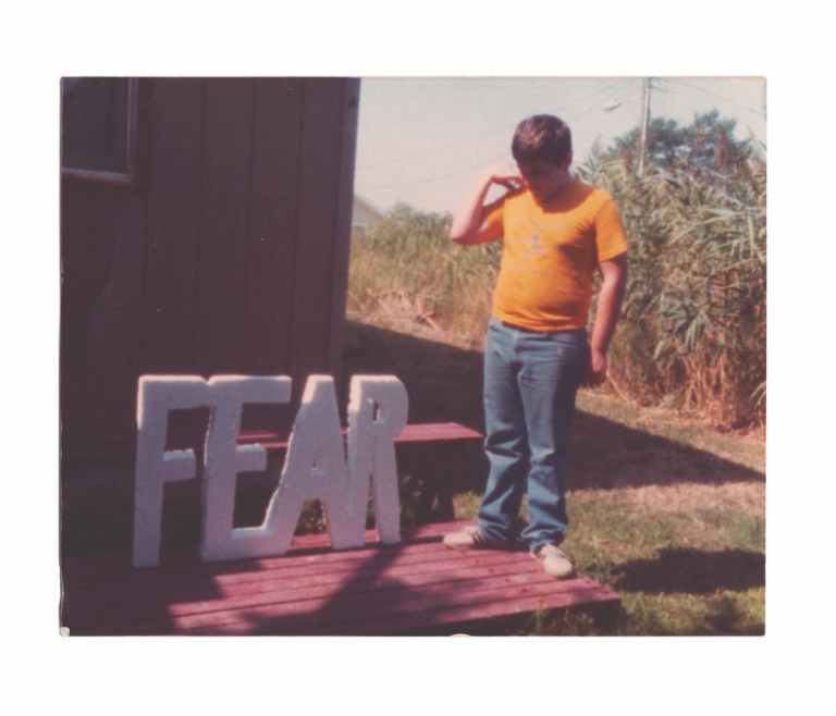 a photograph of a young person wearing a yellow shirt and jeans standing with head turned down looking at a white foam cutout of the letters 'fear' in capitals. there are bushes and the corner of a house behind