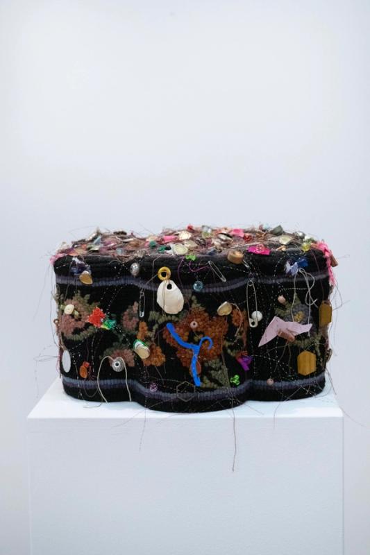 A textile art piece featuring a variety of materials including fabric and found objects, displayed on a white stand.