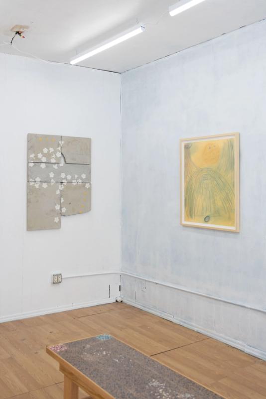 a view of a corner section of a well lit gallery space. three artworks are visible. at foreground is a large bench with dark and very intricate pebbly texture. behind and left is a concrete cast sculpture in mostly gray with white flowers. at right is a framed artwork thats largely yellow with hazy blue sections.