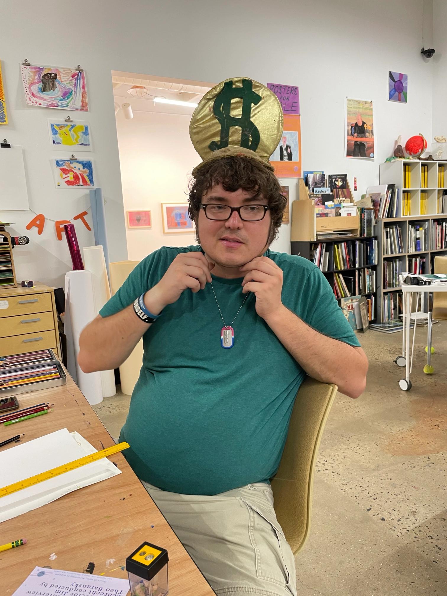 a white skinned man wearing glasses and with angreen shirt and brown hair half smiles at the camera. hes wearing a tall theatrical hat thats gold and has a money sign in green.