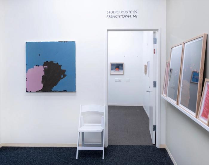 a view into a room through a doorway. at left on the wall is an abstract painting thats mostly light dusty blue with two shapes against each other at the bottom. on left is a light pink shape and at right is a dark brown shape. together they form a loosely round form. at right of the door is a shelf extending into the foreground and in the background another artwork is installed on a wall.
