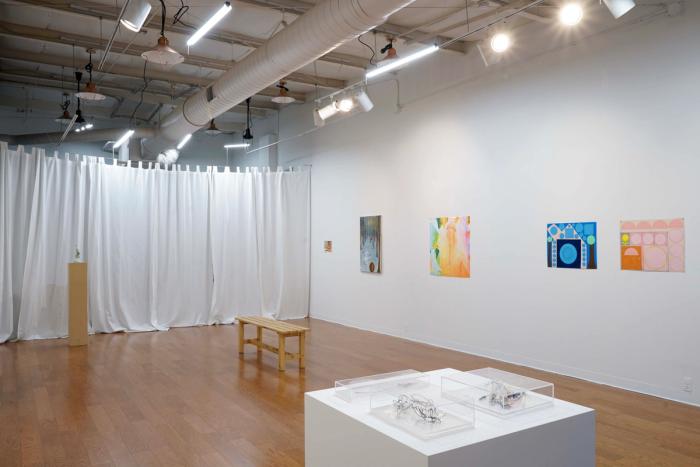 a view of a gallery. there are two dimensional artworks on the walls and a pedestal with a sculpture in the foreground.  the room is white and brightly lit.