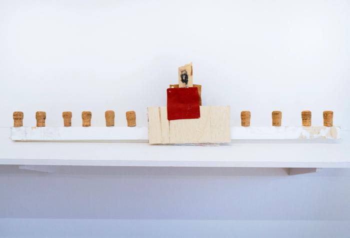 A sculpture titled '‘Building’ Christian Turner, 2024' featuring a wooden structure with a figure, corks, and leather details, mounted on a white shelf.