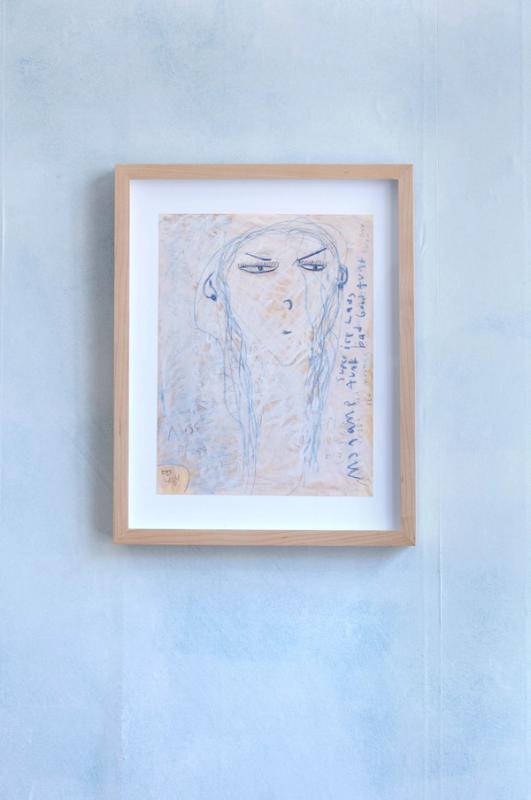 a drawing in a light wood frame against a cloudy blue/gray wall. its of a figure with the head at the top of the page. she has long hair and very suspicious expression as though she's mad. its mostly blue lines smudged with white and on a light tan page.