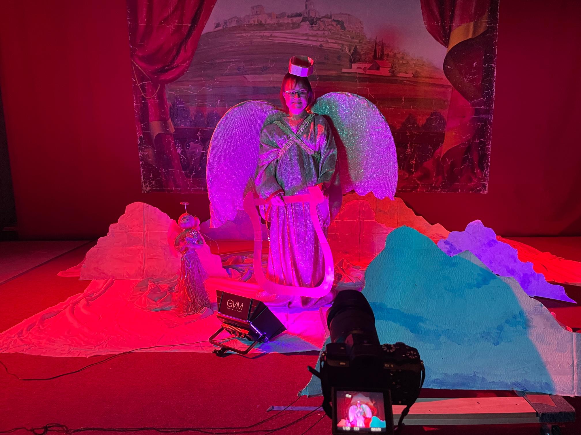 a woman smiles at the camera from a stage. it is lit with bright purples and reds and she wears a golden angel costume with long robe and wings and halo. around her are cardboard cut out clouds and in the foreground a camera points at her making a video
