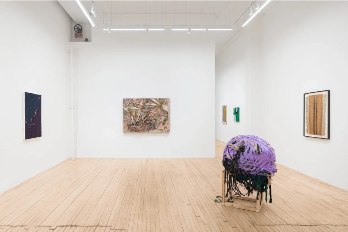 seven artworks are in this photograph of a brightly lit white gallery room with wooden floors. most visible is a large round sculpture which is largely light purple and is perched on a wooden frame, it is made of wrapped ribbon or rope of some kind. the other works are not very discernible at this distance