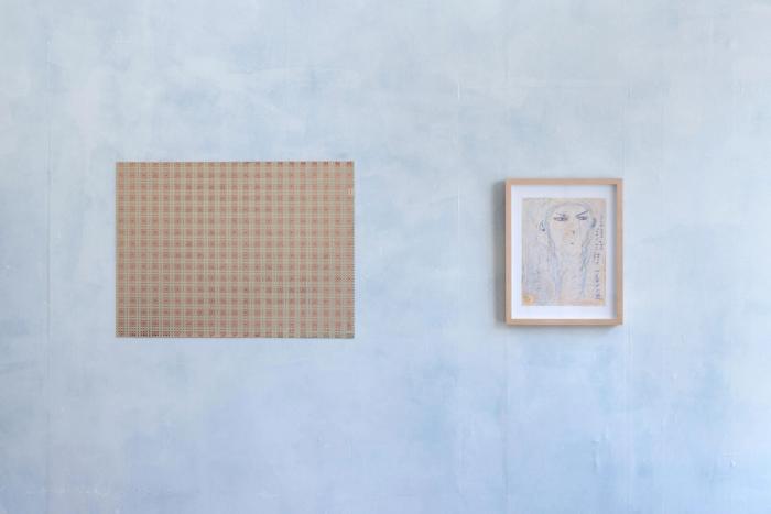 two artworks installed on a cloudy blue/gray wall. on left is a rectangular painting with a stark pink and greenish grid that appears like a textile. to the right is a light-wood-framed artwork with a drawing of a figure's head with long hair in light blue. 