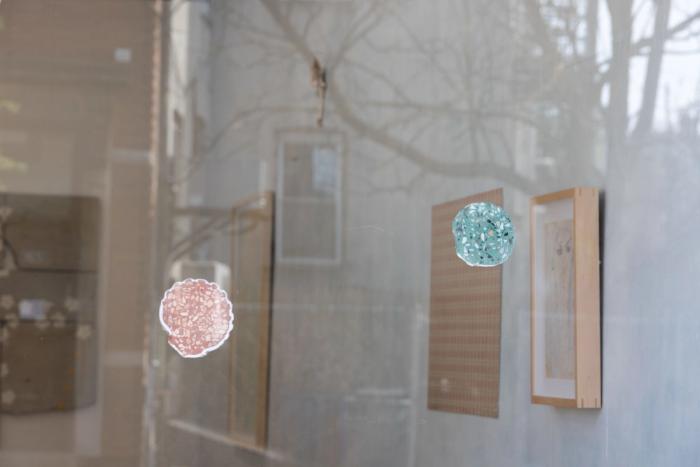 two shell shaped objects are pressed against a reflective glass surface. each one is made of a particulate material with colored fine ground and multicolored pebbles all ground flat. behind is a gallery space with out of focus artworks visible and theres a reflection of tree branches on the glass.