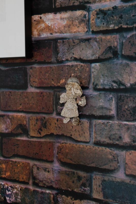 a small sculpture made of coral in the shape of an angel against a vinyl wall printed to look like brick. the angel has broad, simplified features.