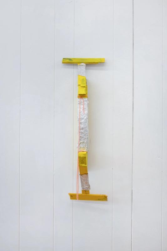 a yellow sculpture installed on a white bead board wall. its mostly yellow and roughly symmetrical. the center section is wrapped in aluminum foil and it loosely takes the shape of a bridge. it has an orange string attached to it and is intalled with the bottom of the bridge against the wall