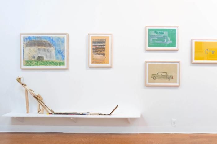 a white gallery wall with five drawings in frames hanging. they are all of different things, a house, a pattern,  a car are discernible. below them is a low shelf about 10 inches from the ground with a long sculpture on it made of wood and cardboard scraps